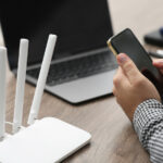 A man uses a smartphone next to a laptop and router.