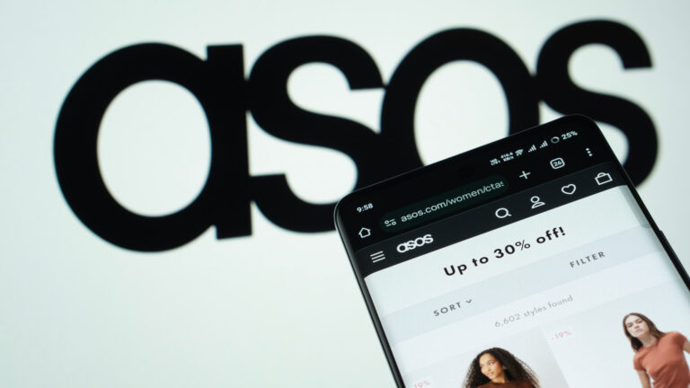 Photo of a smartphone open to the Asos website. The retailer's logo is displayed in black on a slightly off-focus white backdrop behind the phone.