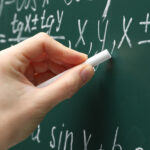 A hand with chalk calculating math problems on a classic green chalkboard.