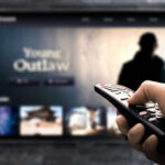 TV out of focus in the background, with foregrounded hand holding a remote pointed at it.