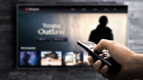TV out of focus in the background, with foregrounded hand holding a remote pointed at it.