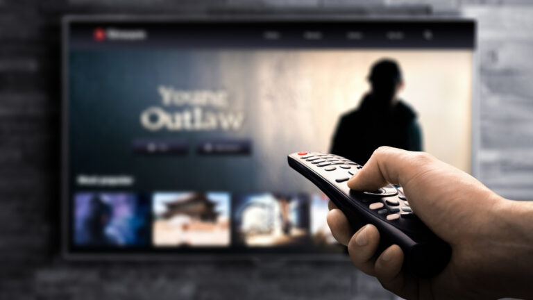 TV out of focus in the background, with foregrounded hand holding a remote pointed at it.