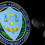 Photo of a black screen displaying the insignia for the US Federal Trade Commission. Two fingers near the image are faintly illuminated by the ambient light.